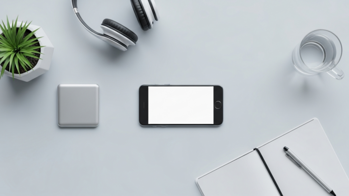 Flat-lay desk setup for testing AI girlfriend chat apps: a blank-screen smartphone, headphones, an open notebook and pen, a glass of water and a small potted plant